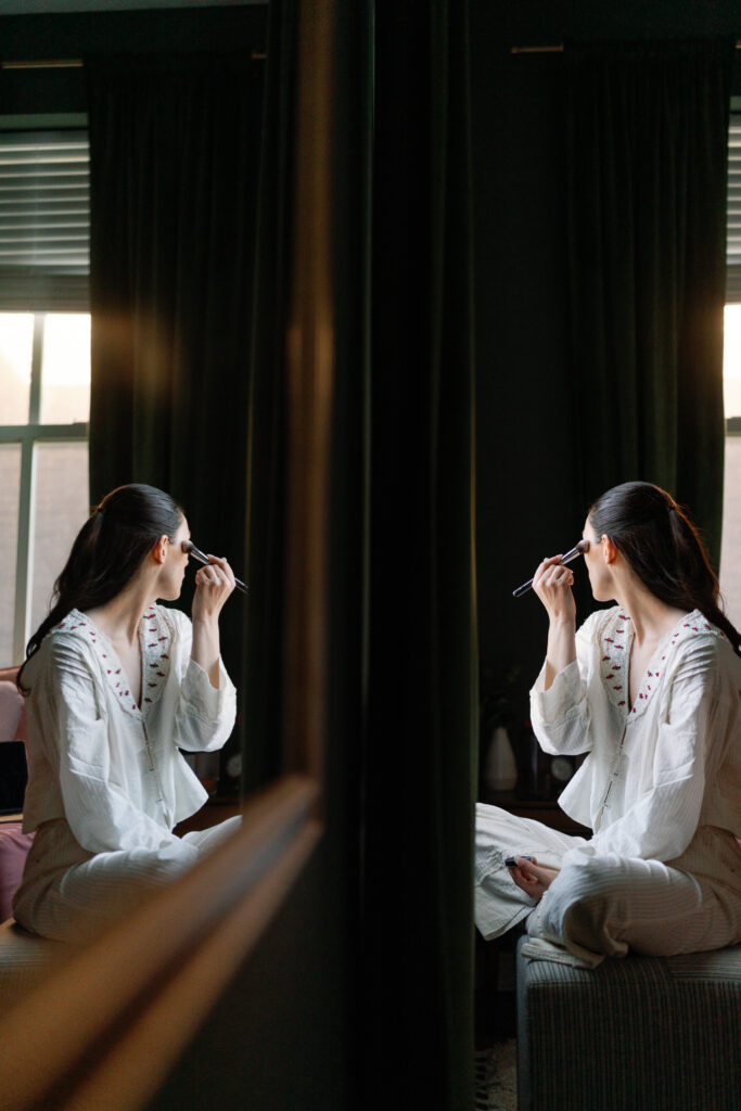 bride doing her makeup sitting by a window with a reflection off a mirror that creates a unique image