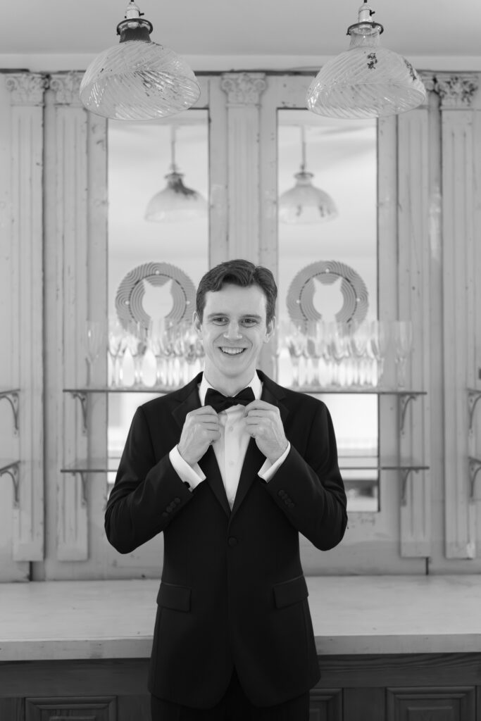 black and white photo of a groom in a tuxedo adjusting his bow tie and laughing