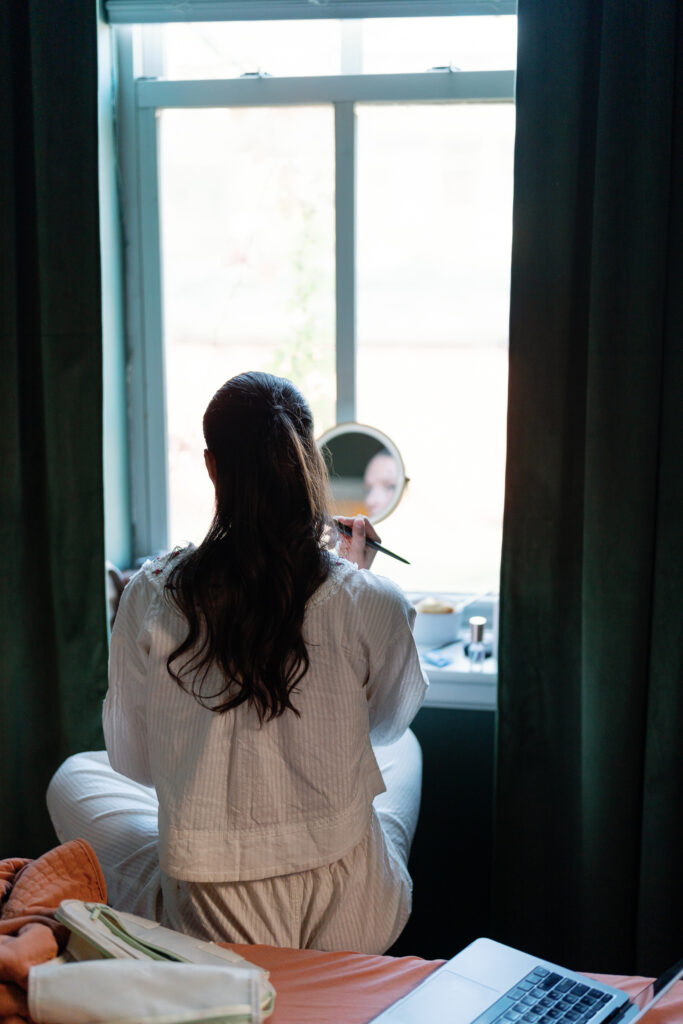 Bride sitting at a window doing her makeup using the light from the window