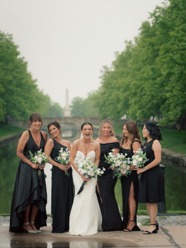 Bride laughing with bridesmaids in black dresses beside canal at The Marquee at New Town