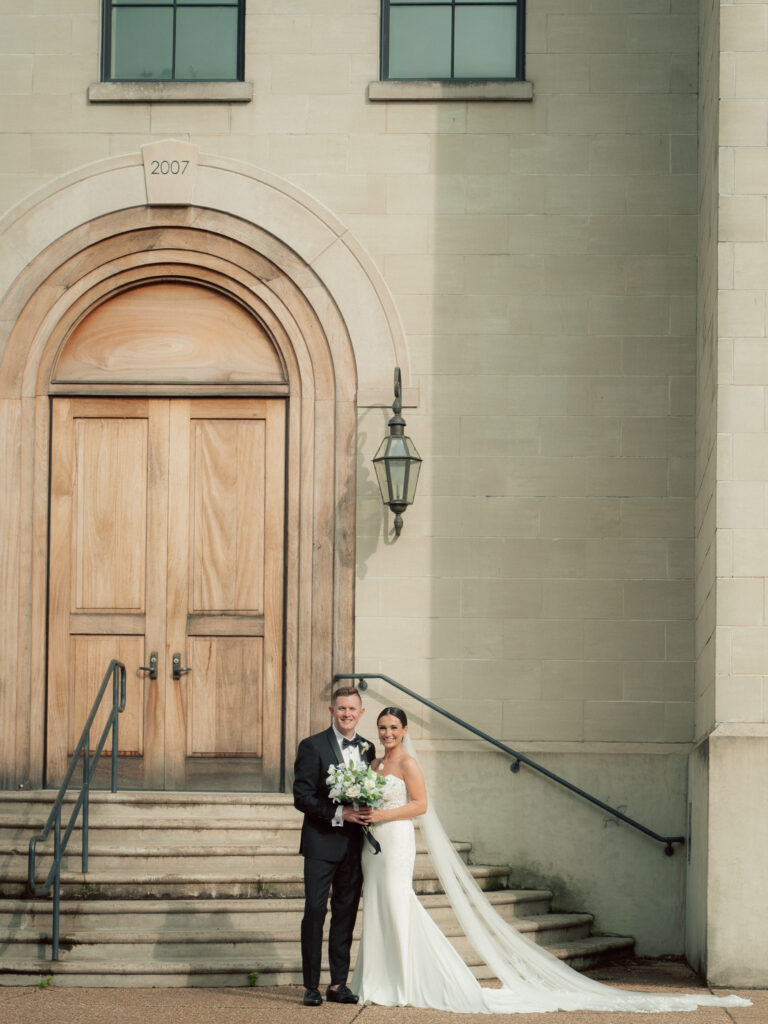 Bride and groom standing on church steps beneath arched wooden door at The Marquee at New Town