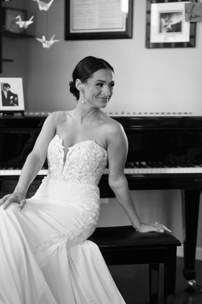 Bride in elegant wedding dress smiling while seated at piano in New Town home before ceremony