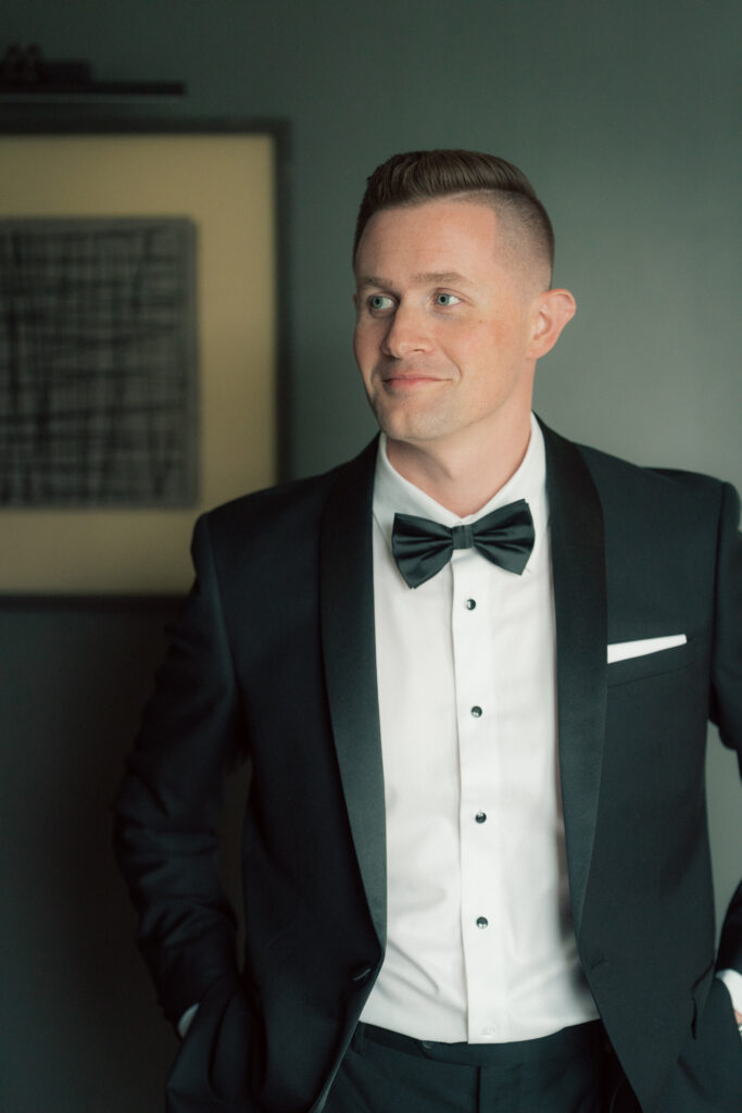 Groom in classic black tuxedo with bow tie standing in soft natural light before wedding at The Marquee at New Town