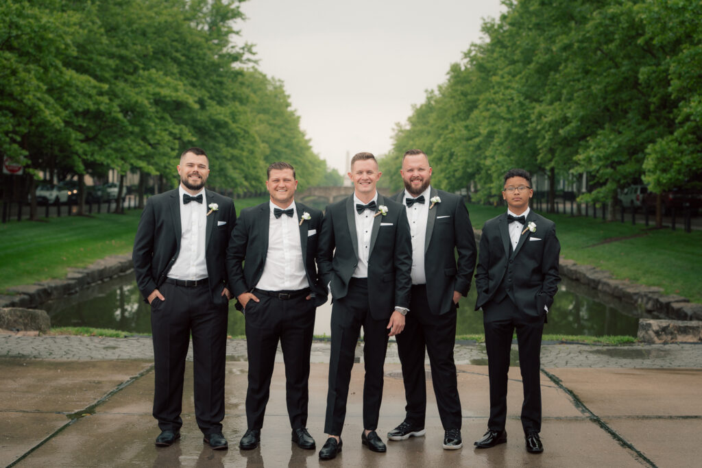 Groom standing with groomsmen in black tuxedos in front of canal at The Marquee at New Town
